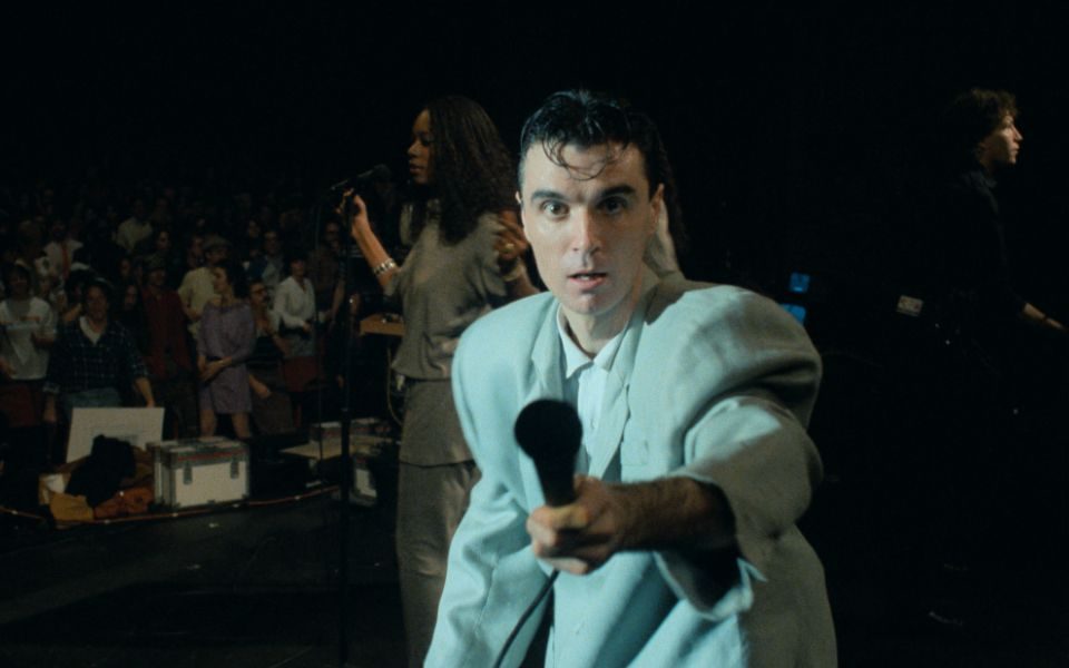 On stage in an oversized grey suit, David Byrne holds up a microphone to the camera. A singer and crowd are visible in the background.