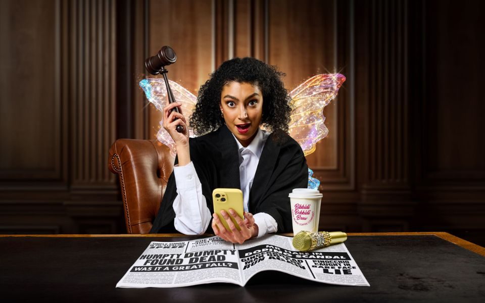 A woman in a judge's outfit and sparkling fairy wings, holding a gavel in one hand and a smartphone in the other. In a courtroom, she is sat at a desk with a newspaper, coffee cup and golden microphone.