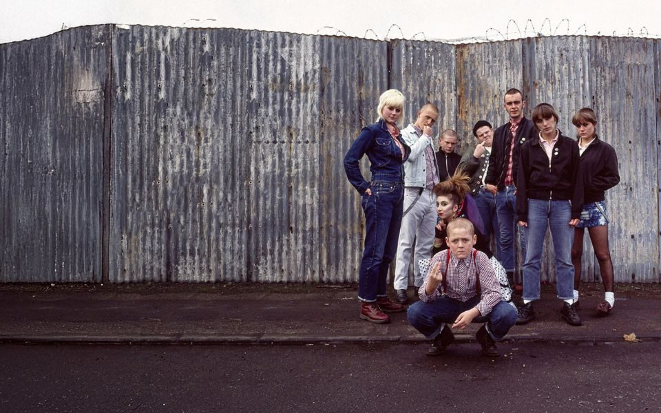 A group of skinheads and punks in 1970s clothing, stood on a street in front of a corrugated metal fence. In the front, a young boy with a shaved head and red braces crouches down at gestures towards the camera.