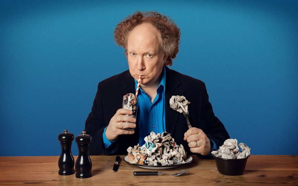 Andy Zaltzman sits at a wooden table, sipping from a glass filled with scrunched up newspaper. In front of him there is a plate, cutlery and bowl also filled with discarded newspaper.