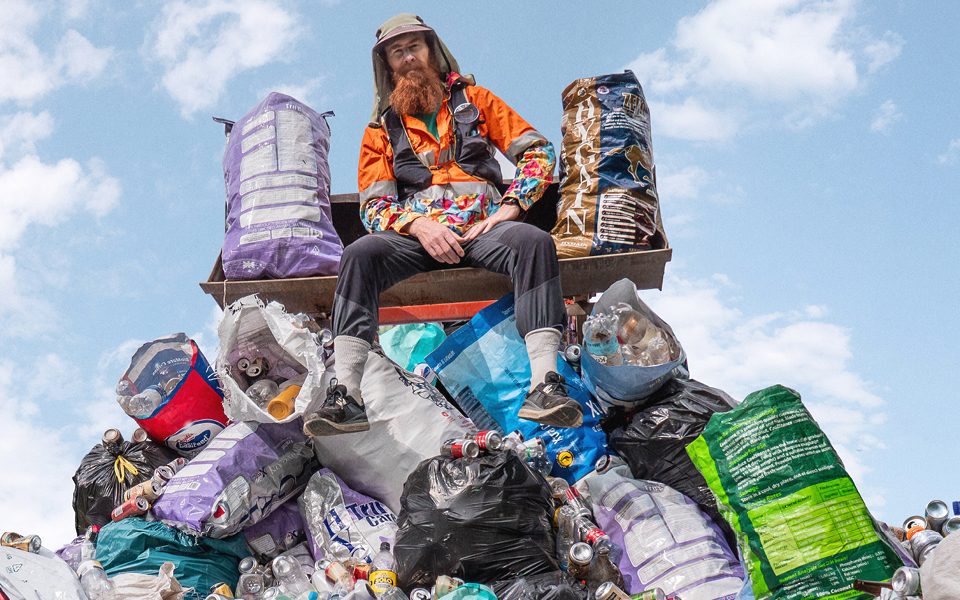 A man with a long ginger beard, a floral shirt and orange high-vis jacket sits on top of a small mountain of bin bags containing cans and plastic bottles in front of a cloudy sky.