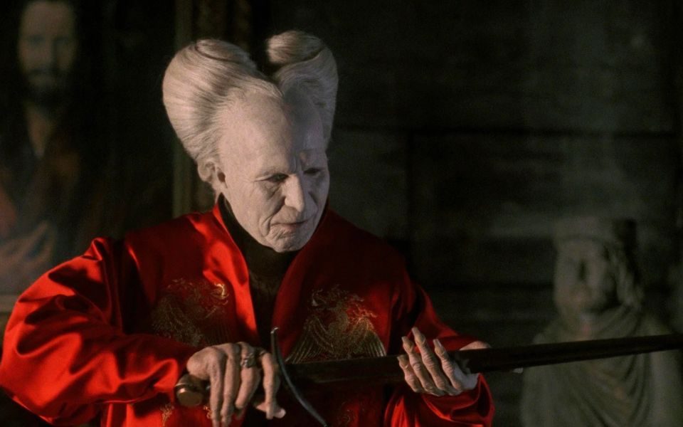 A man with white dramatically-styled hair and pale skin looks down at a sword in his hands.