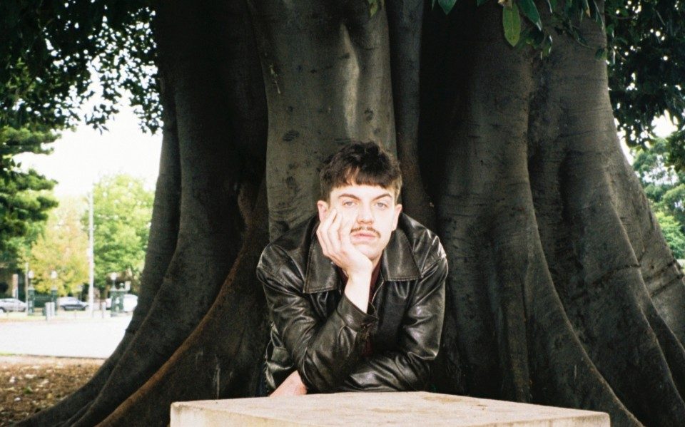A man in a leather jacket rests his head in his palm, his arms folded across a white concrete block in a forest.