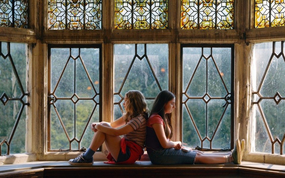 Two girls sit back-to-back on a windowsill in front of stained glass windows.