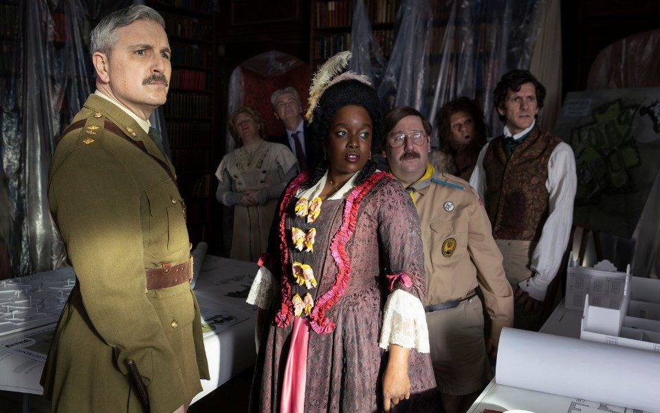 A woman in a regal period dress stands beside a moustached man in a scouts uniform, and a man in a green army uniform. In the background are more people in period clothing in a library covered with clear tarpaulin sheets.