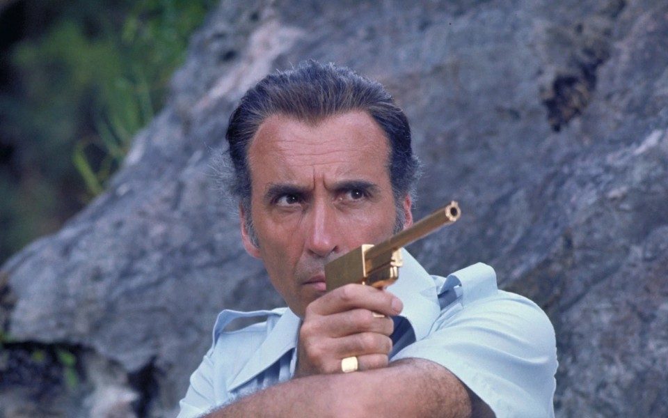 With a stern expression, a man by a rock holds a golden gun and points it off-camera.