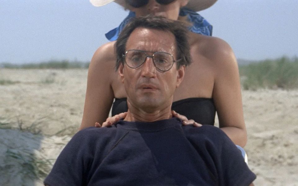 A man gazes intensely toward the camera. In the background, a woman and a beach.