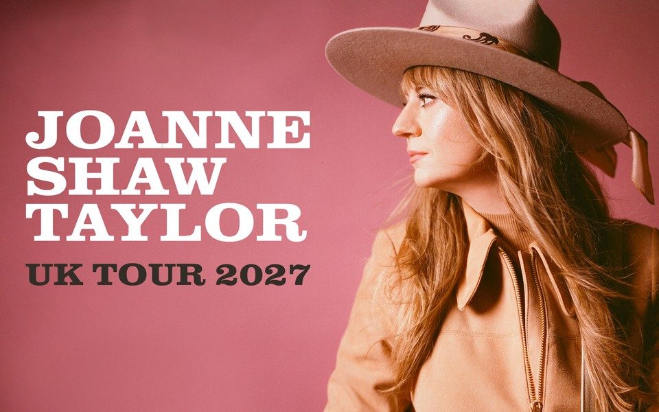 Joanne Shaw Taylor looks off to the side, holding a well-worn acoustic guitar and cowboy hat against a pink backdrop. Text reads: Joanne Shaw Taylor. UK tour 2027.
