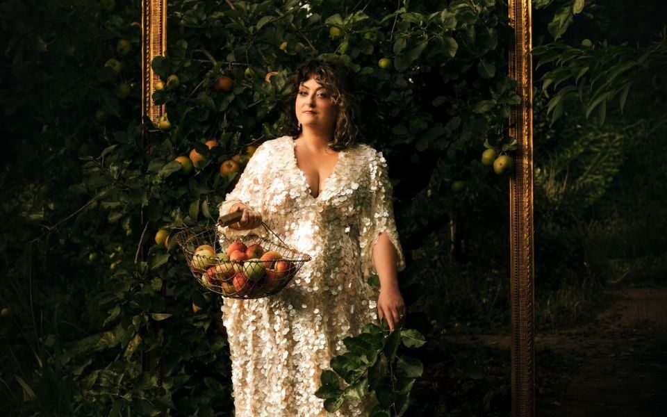 Kiri Pritchard-McLean wearing a long sequined dress, holding a basket of apples, in front of a golden photo frame containing the branches of a large apple tree.