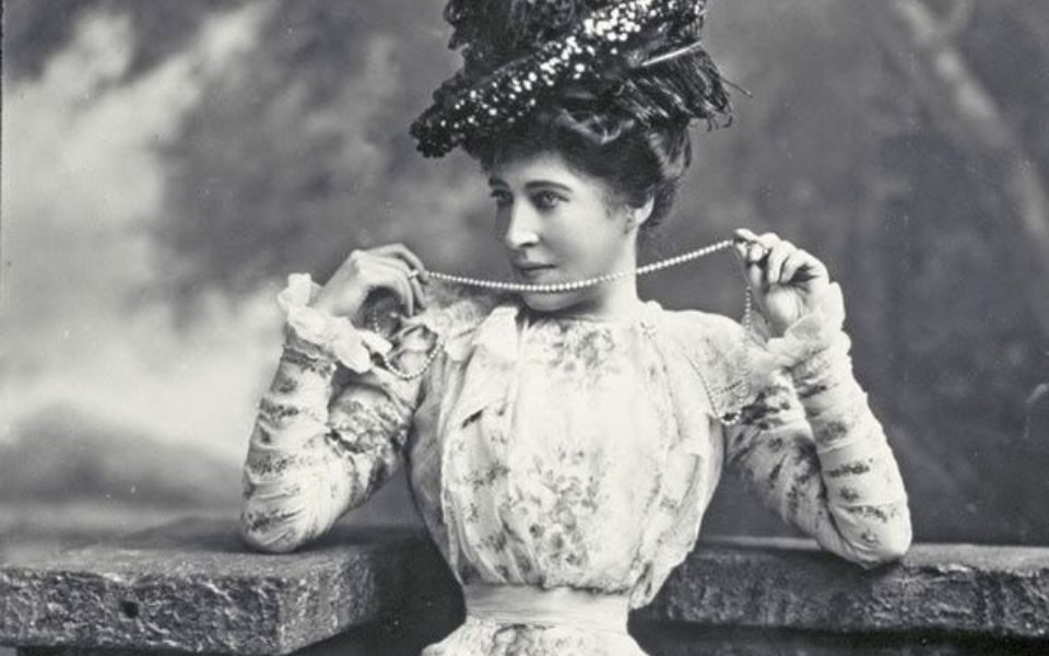A black and white image of a woman in a fascinator and corseted dress, holding up a pearl necklace