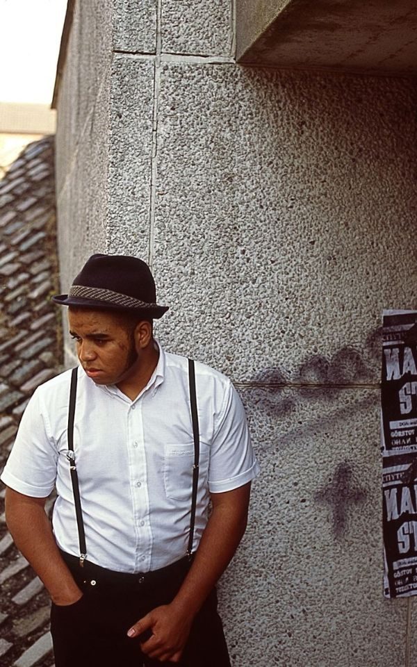 A young man stands beside a concrete wall adorned with gig posters and graffiti. He wears a pork pie hat, white shirt and braces.