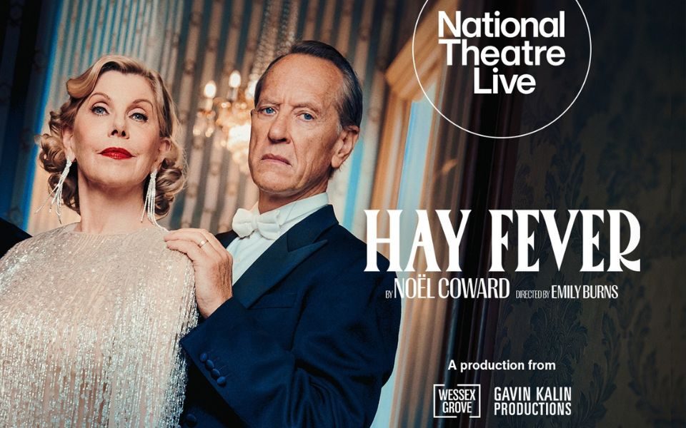 With serious expressions, Christine Baranski wears dangly earrings and a shimmering dress, standing beside Richard E Grant in a dark tuxedo with a white bowtie. A chandelier is visible by a window and patterned wallpaper behind them. Text reads: National Theatre Live. Hay Fever by Noel Coward. Directed by Emily Burns. A production from Wessex Grove and Gavin Kalin Productions.