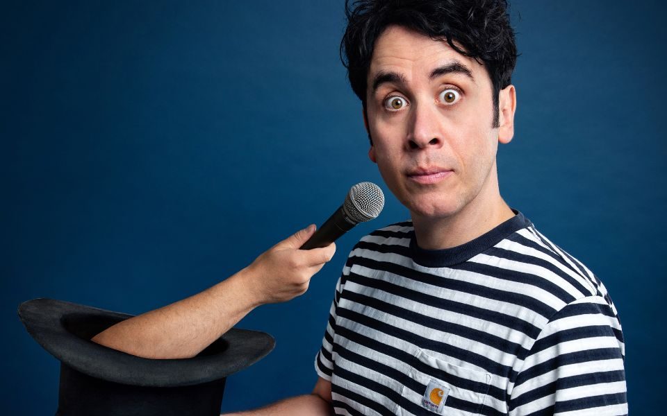 In a striped t-shirt, Pete Firman displays a shocked expression to the camera as an arm holding a microphone emerges from a top hat he is holding.