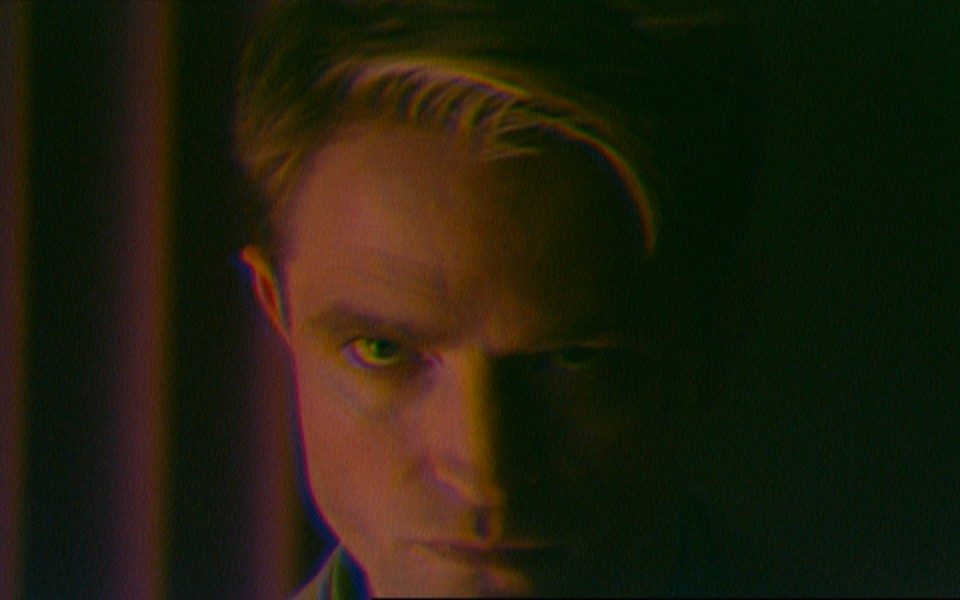 On a grainy digital screen, a man looks intently into the camera, his head tilted down and half in shadow.