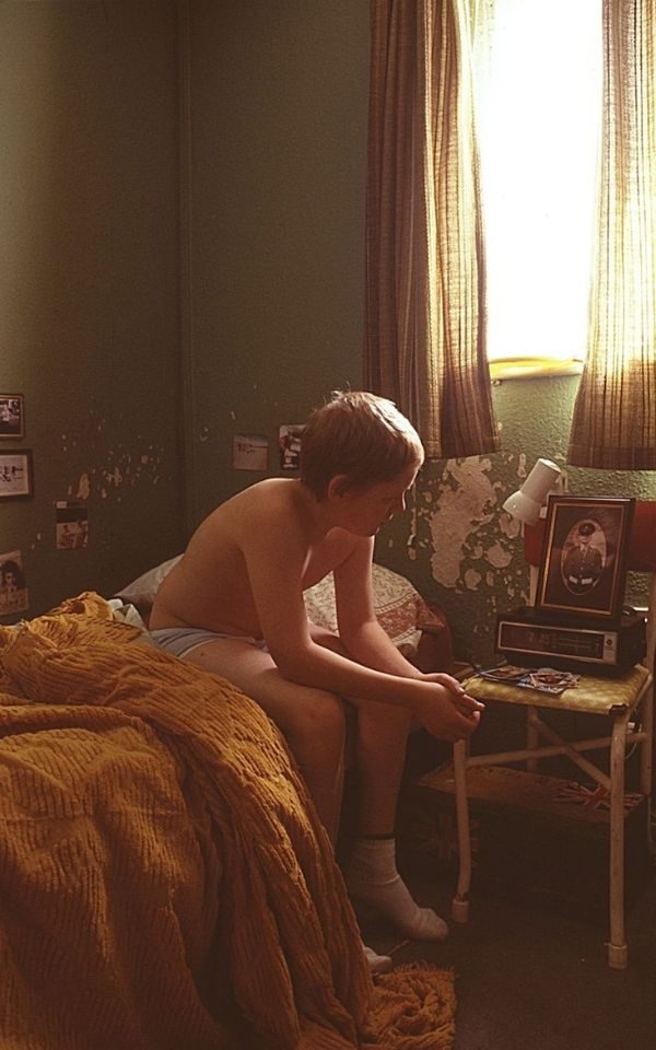In a bedroom with peeling paint and photos on the walls, a young boy in underwear sits on a bed, looking at a framed picture beside him.