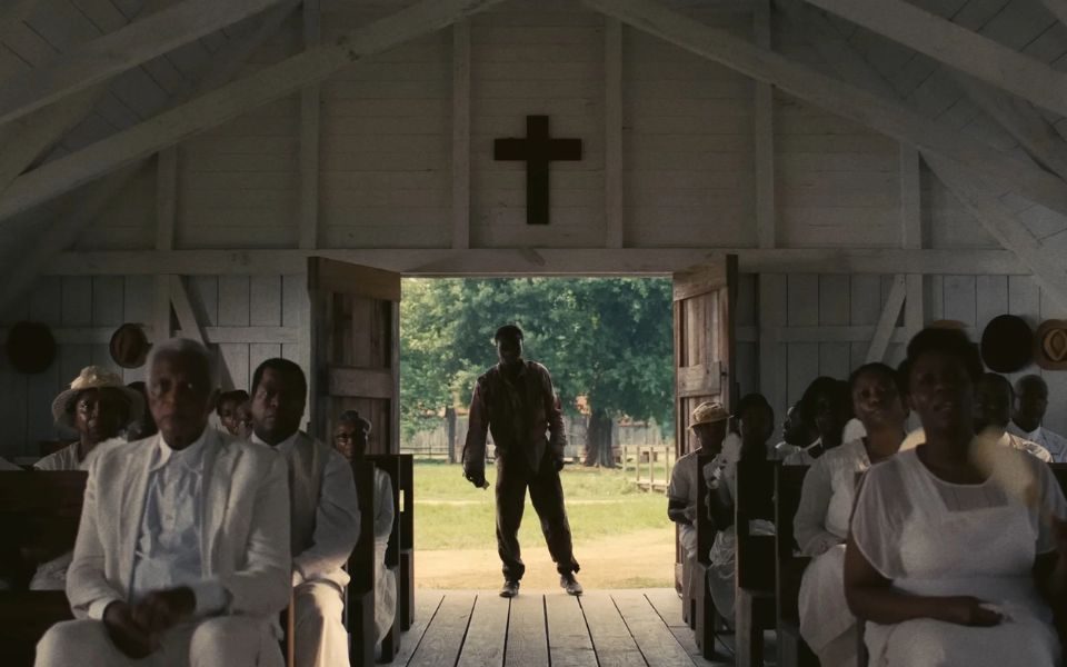 A congregation of churchgoers sat in the pews of a church wearing white. In the background, a figure limping through the door in silhouette.