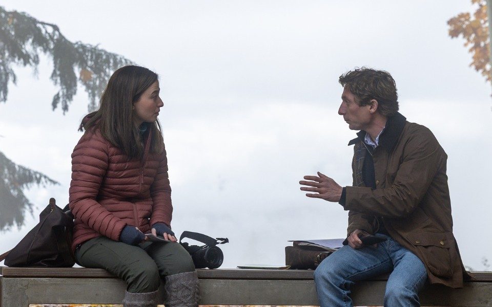 In front of a snowy backdrop, two people sit on a bench having a conversation.