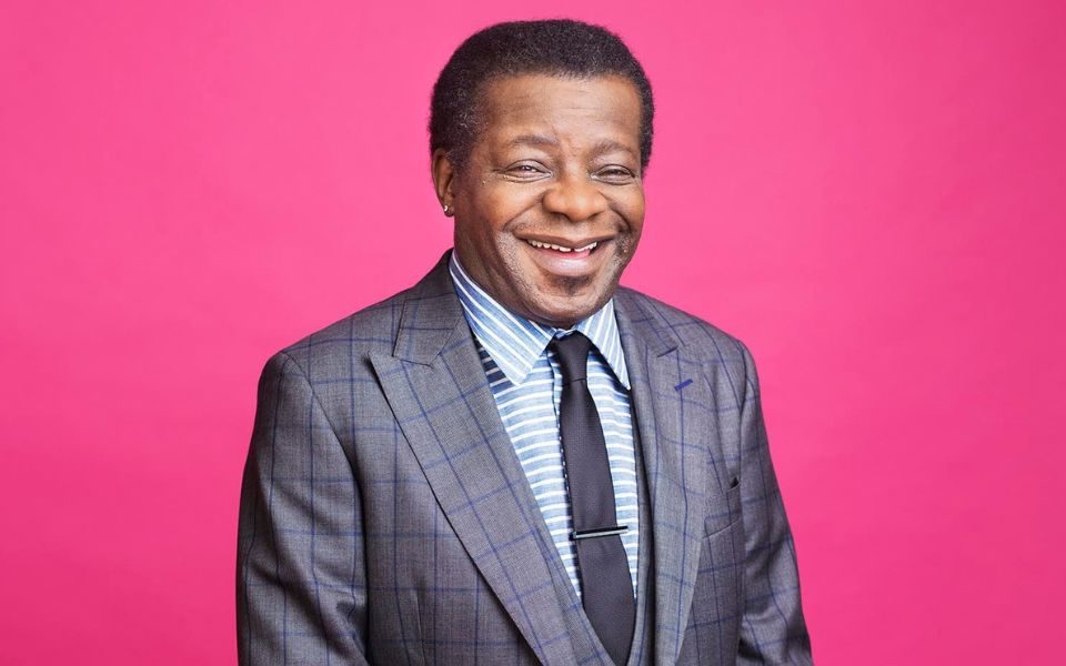 Wearing a grey suit in front of a pink background, Stephen K Amos stands laughing.