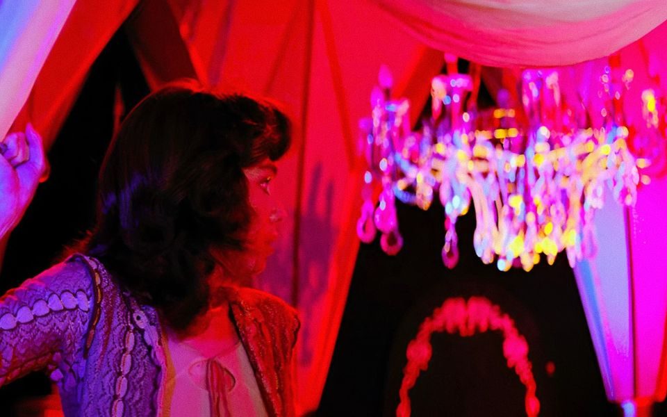 In bright technicolour, a woman in the foreground looks behind her. A chandelier is visible in the background.