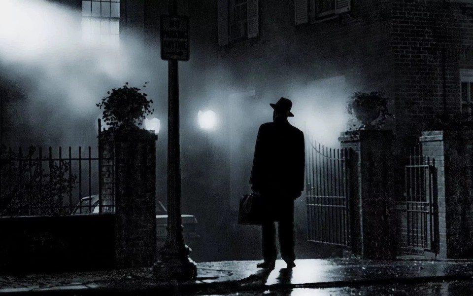 Silhouette of a man in a hat holding a briefcase outside a house on a dark, foggy street.