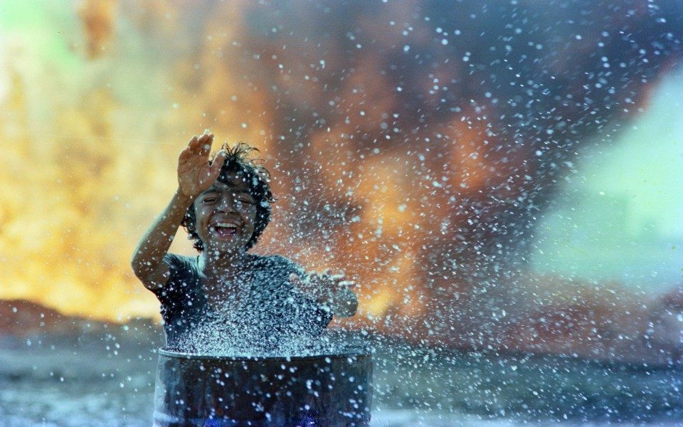 A young boy playfully bangs his hands on top of a barrel, causing water to spray in the foreground. Smoke and fire fill the background.