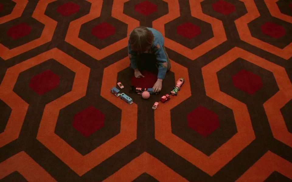 On a carpet patterned with large red-and-orange hexagons, a child plays with toy vehicles.