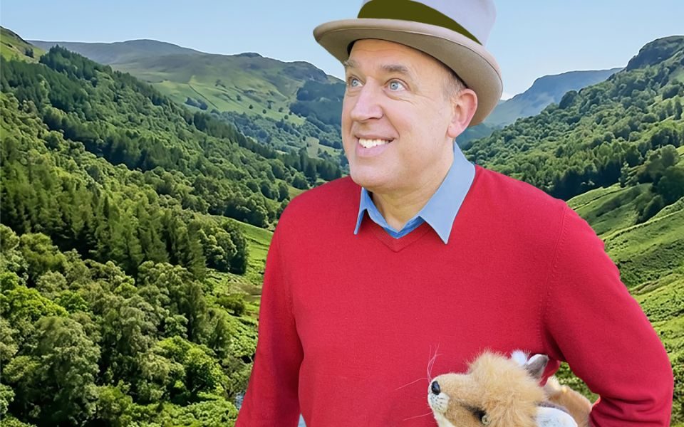 Stood smiling in front of a green valley, Tim Vine wears a red jumper and top hat, holding a fluffy toy fox.