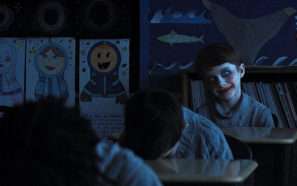 In a dark classroom, children sit with their head against their desks. At the back of the room, a child smiles wide-eyed wearing messy makeup.