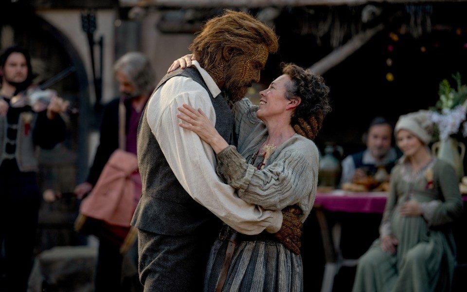 A man made of wicker and a woman hold each other closely and smile amongst a crowd of people in period costumes.